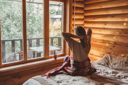 Frau in warmer Decke entspannt auf gemütlichem Bett in Blockhütte im Winter.